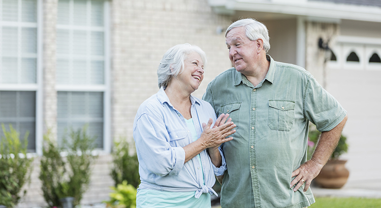 A senior couple in their 60s standing together in the front yard, in front of their house. They are retired homeowners, smiling and looking at each other
